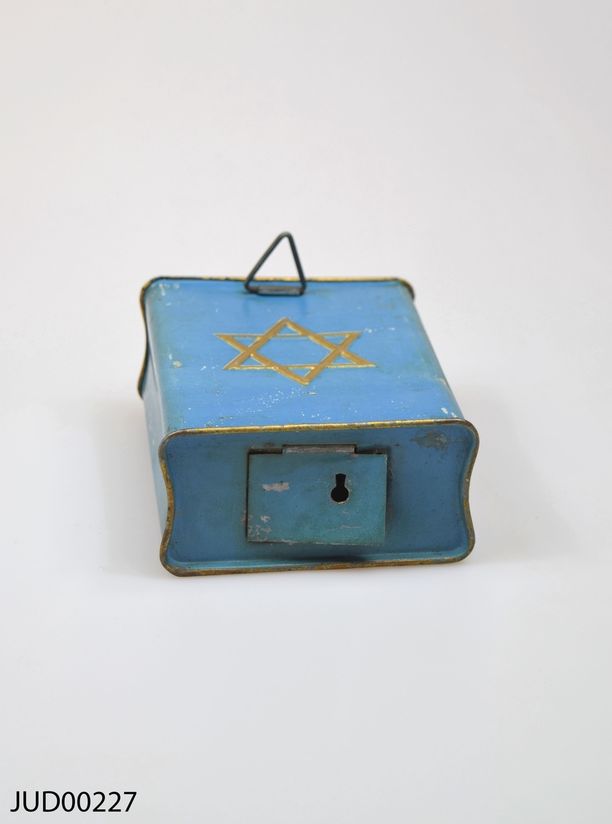Insamlingsbössa för KKL, tillverkad av plåt. Bössan är målad blå, med en gul Davids-stjärna på framsidan samt hebreisk text. "J.F".