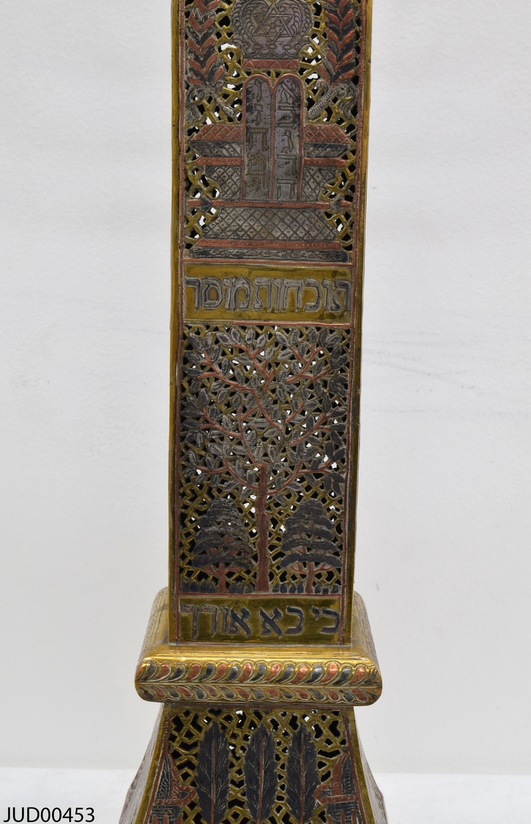 Stor chanukkaljusstake tillverkad av mässing, i form av en obelisk dekorerad genombruten dekor i rött och svart. Davidsstjärna på toppen mellan ljushållarna.