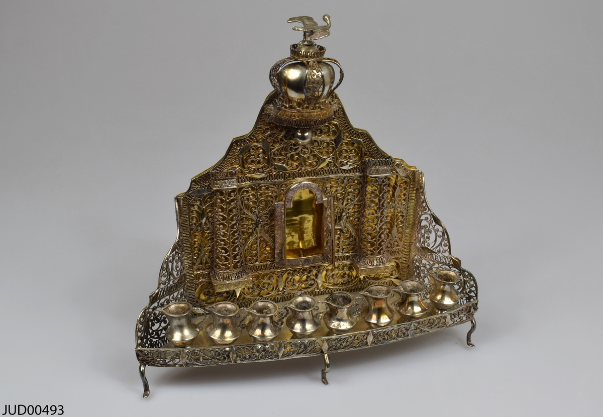 Chanukaljusstake tillverkad av silver, med åtta kannor som ljushållare. Två lösa flankerande ljushållare. Dekorerad med genombruten filigran, samt ett torahkabinett, en krona och en fågel. 

Tillverkad i Baal Shem Tov-stil.