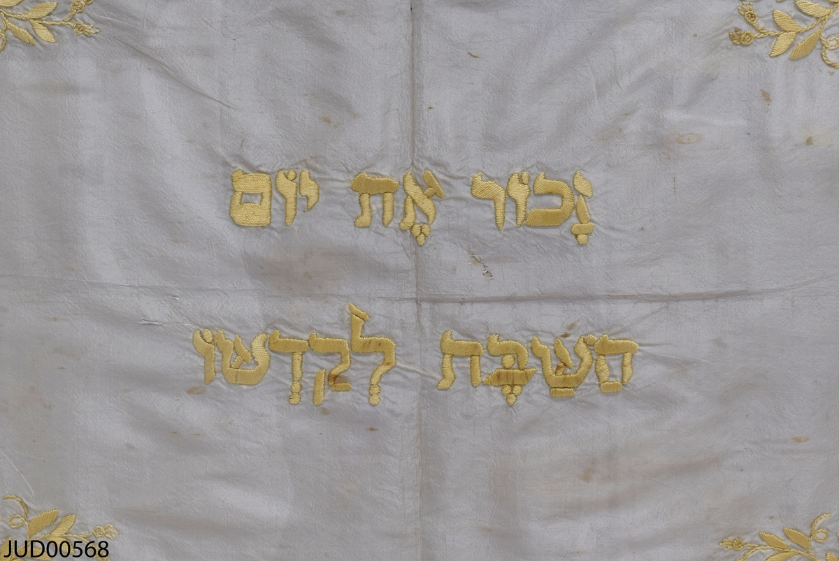 Challaduk tilllverkad av vitt siden, med ljusgula fransar runt kanten. Broderi i form av blad i samma gula nyans i varje hörn. I mitten broderade hebreisk text. Monterad i glas och guldfärgad ram.