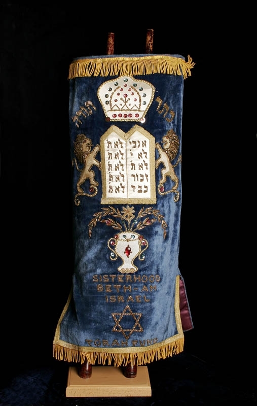 Torahrulle med tillhörande torahmantel. Torahmanteln tillverkad av blå sammet och dekorerad med guldbroderier i form av texten "Sisterhood Beth Israel" samt en vas med halvädelstenar. Själva torahrullen är tillverkad av pergament.