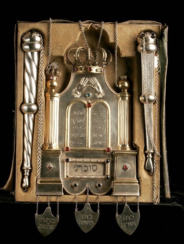 Torahsköld med två torahpekare liggandes i låda. Lådan är klädd med svart skinn på utsidan och beige sammet på insidan. 

Skölden är tillverkad av silver och dekorerad med färgade stenar. Kolonner på sidorna och krona på toppen, samt tre hängande plaketter i nederkant. 

Pekarna är tillverkade av silver. En är vriden och utan annan dekor, medan den andra har en rak fyrkantig form och dekorerad med ristad hebreisk text.