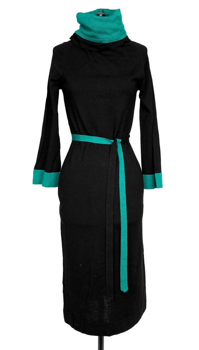 A-formet kjole i strikk med rette ermer. Dobbel nedbrettkrage i grønn utenpå svart. Grønt nederst på ermene. Smalt knytebelte der halvparten er svart og halvparten grønt. 