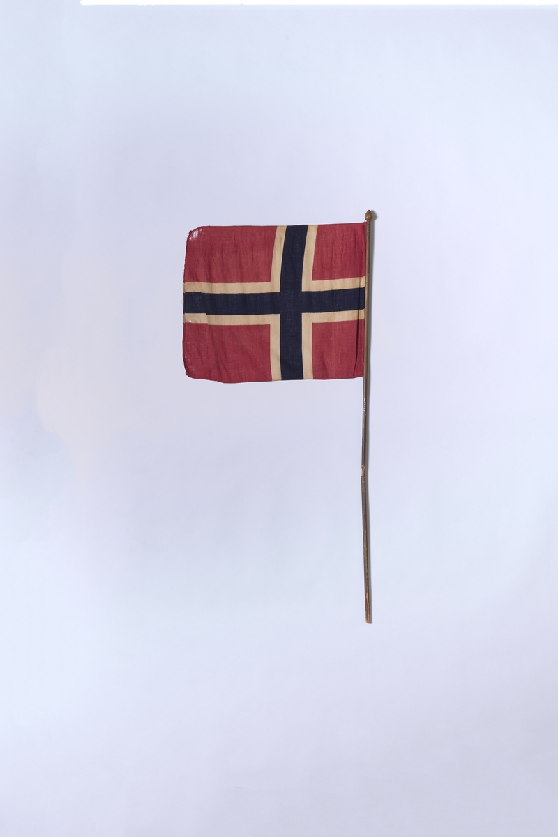 Det norske flagg, montert på trestang. Flagget er i bomull og har trykte farger. Stanga ser ut til å ha brukket for så å bli reparert. 