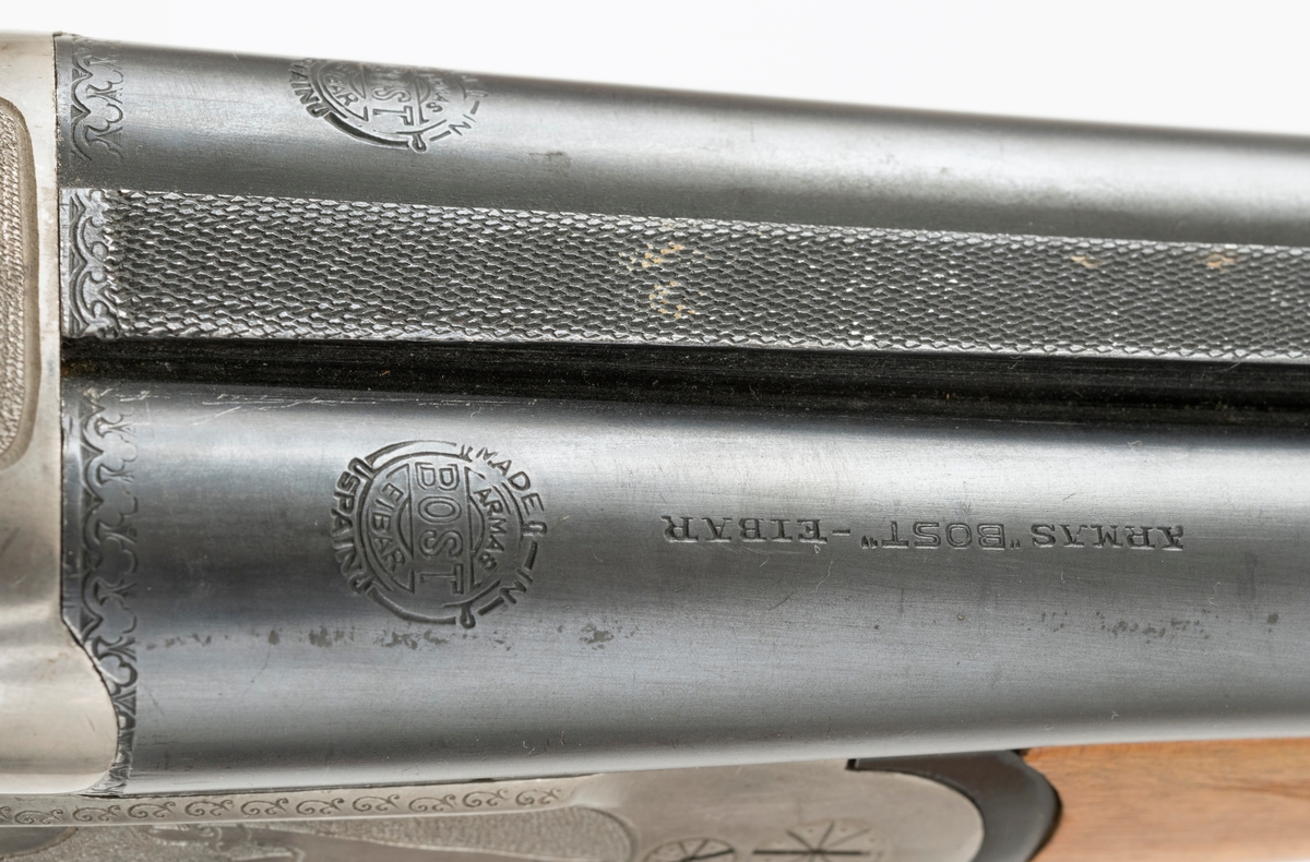 Dobbeltløpet hagle (side by side) for kaliber 20 med bærereim. Det mangler en skrue i skjeftet. Geværet har enkel gravyr med motiv av stående fuglehund.