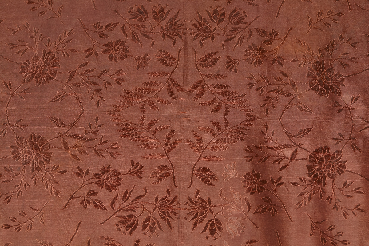 Jacquard-/damaskvevet silkesjal med frynser. Sjalet er høybrunt, og har blomstermønster med bl.a. liljekonvall. Mulig produsent K.A. Almgren, Stockholm (kilde: tidligere registrering)
