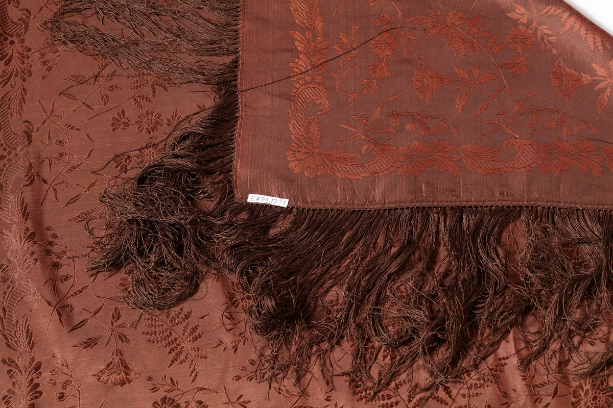 Jacquard-/damaskvevet silkesjal med frynser. Sjalet er høybrunt, og har blomstermønster med bl.a. liljekonvall. Mulig produsent K.A. Almgren, Stockholm (kilde: tidligere registrering)
