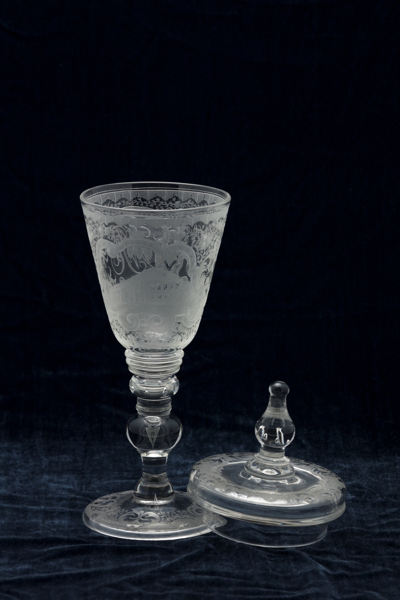 Pokal med lokk, glass. Fot har graverte blomster  og knekkede bånd. På cupa gravert bilde av Bogstad på en side, på den andre: N.W.J.  W.P.E.