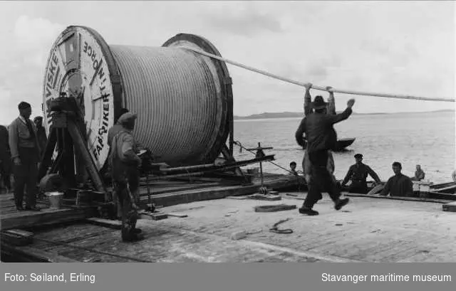 Kablenedlegging utenfor Hundvåg. Arbeider med å legge elektrisk kabel fra båten Rosenberg. Kablerull merket Skandinaviske fabrikker i Norge - Oslo. 
Flere tilskuere og arbeidsfolk i robåter