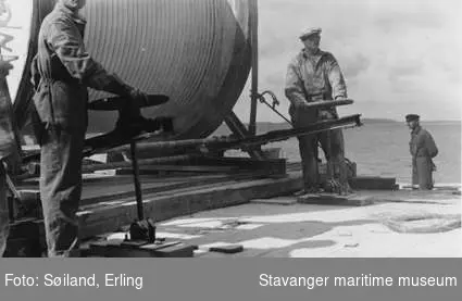 Kablenedlegging utenfor Hundvåg. Arbeider med å legge elektrisk kabel fra båten Rosenberg. Kablerull merket Skandinaviske fabrikker i Norge - Oslo. 
Flere tilskuere og arbeidsfolk i robåter.