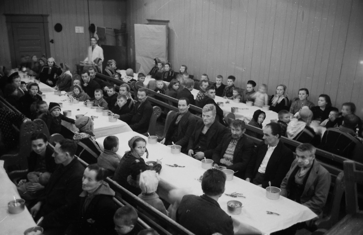 Tvangsevakuerte fra Porsanger sitter samlet til et måltid ved flere langbord og på kirkebenker i Metodistkirken i Trondheim julaften 1944. Mange av dem snur seg mot fotografen.
