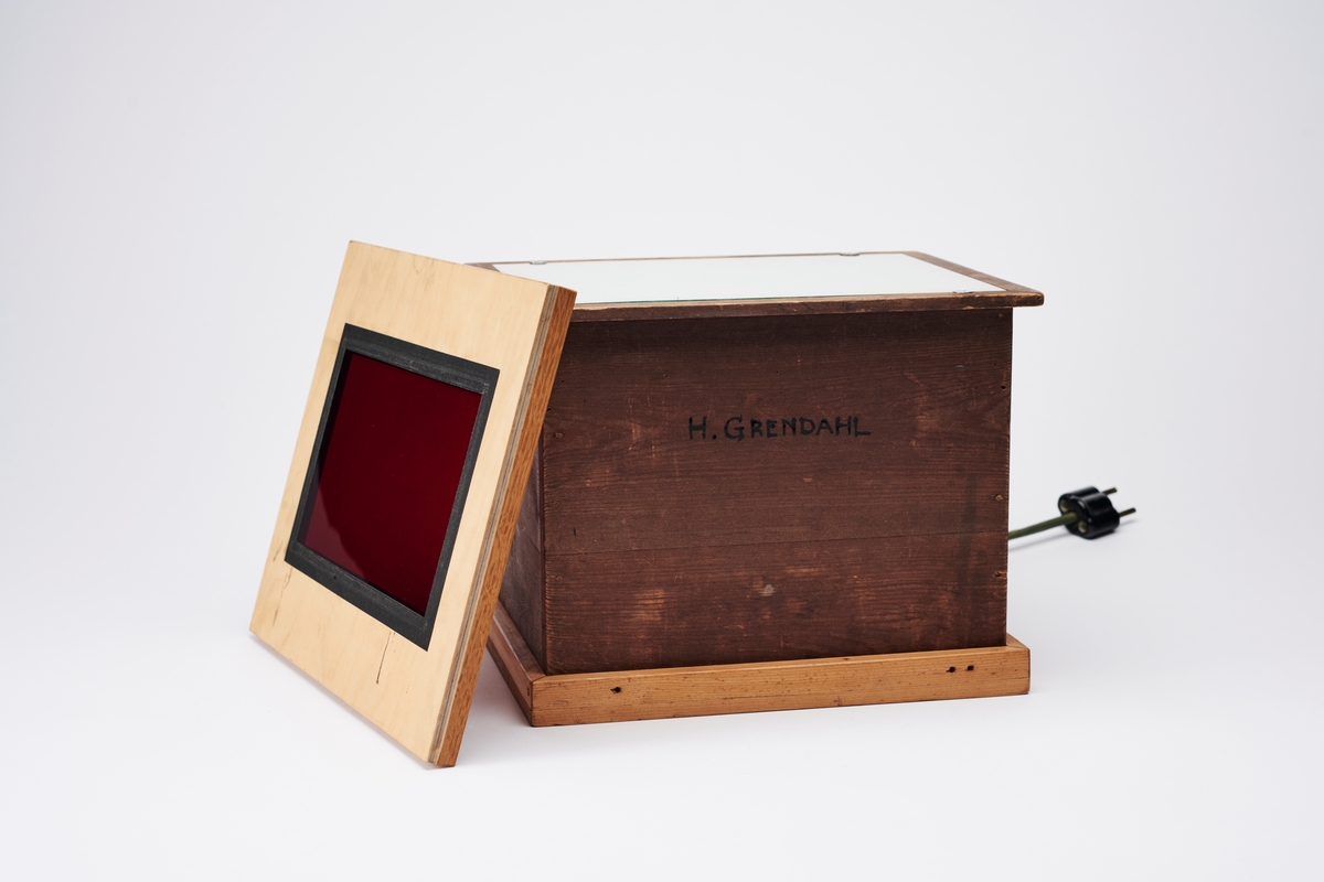 Lyskasse med matt glass. Kassen kan fungere som et mørkeromslys ved bruk av rammen med rødt glass. Konstruert av arkitekt Hans Grendahl (1877-1957).