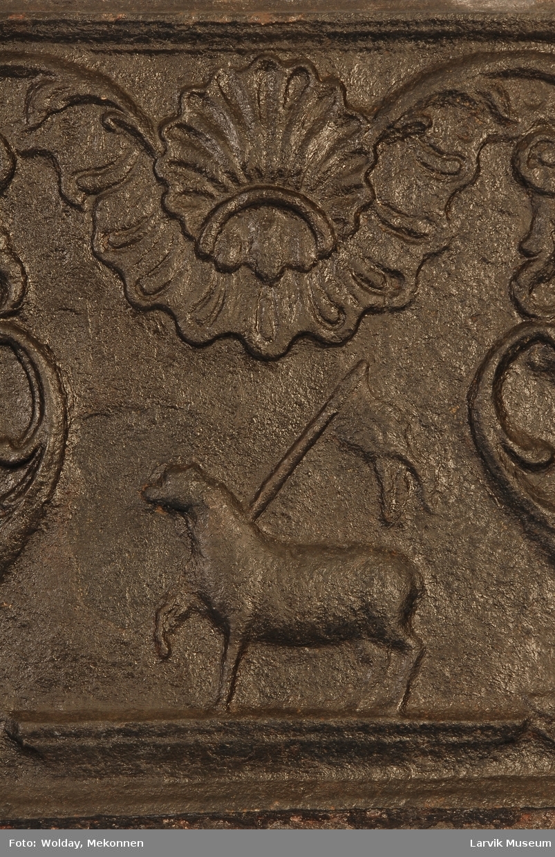 lam m/seiersfane (Kristussymbol) på lavt postament, omgitt av symmetrisk rokokko-ornamentikk...