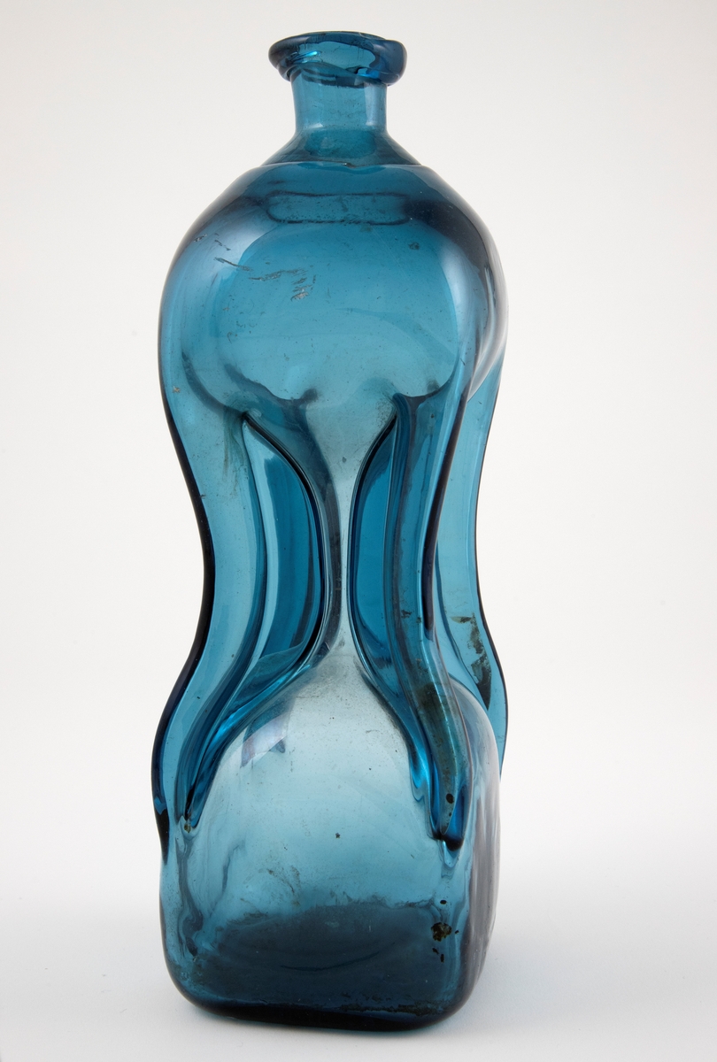 Klukkflaske i gjennomskinnelig mørkeblått glass. Timeglass-lignende utforming, hvor den tilnærmet kvadratiske over- og underdelen er forbundet med et tynt rør. Lav hals med glatt kantring, samt puntemerke på undersiden.