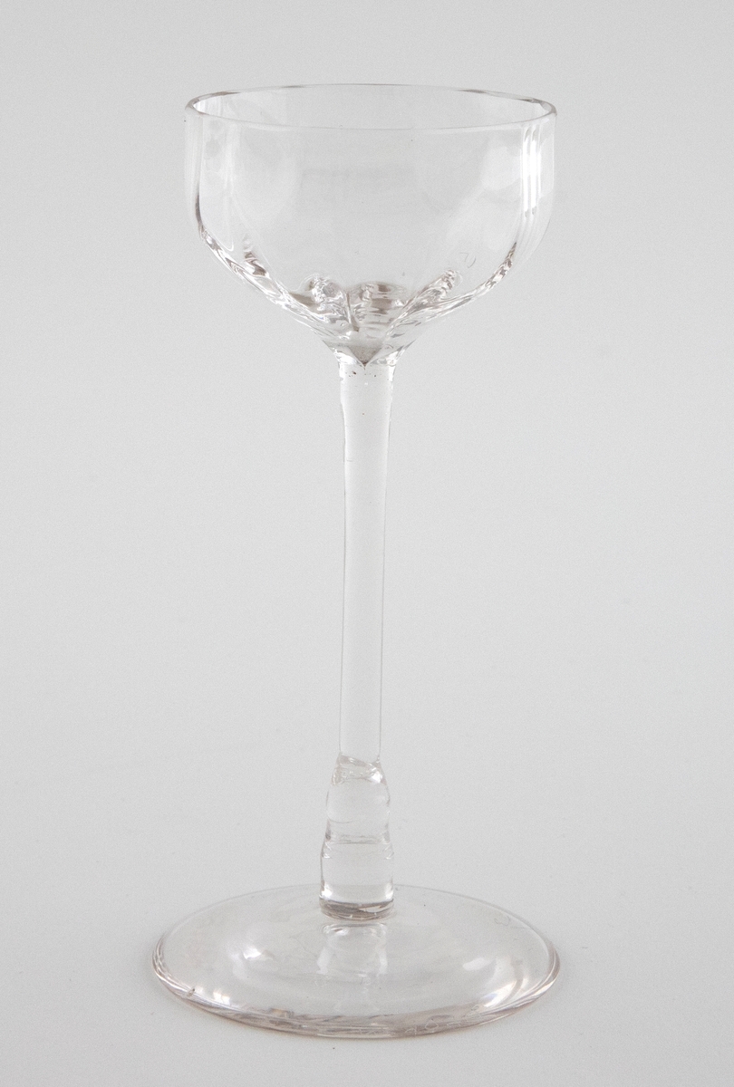 Likørglass i klart glass. Kuleformet lav kupa som er dekorert med punktdekor. Hviler på en slank stett, hvor overgangen til den sirkulære fotplaten er markert med en kuleformet vulst.