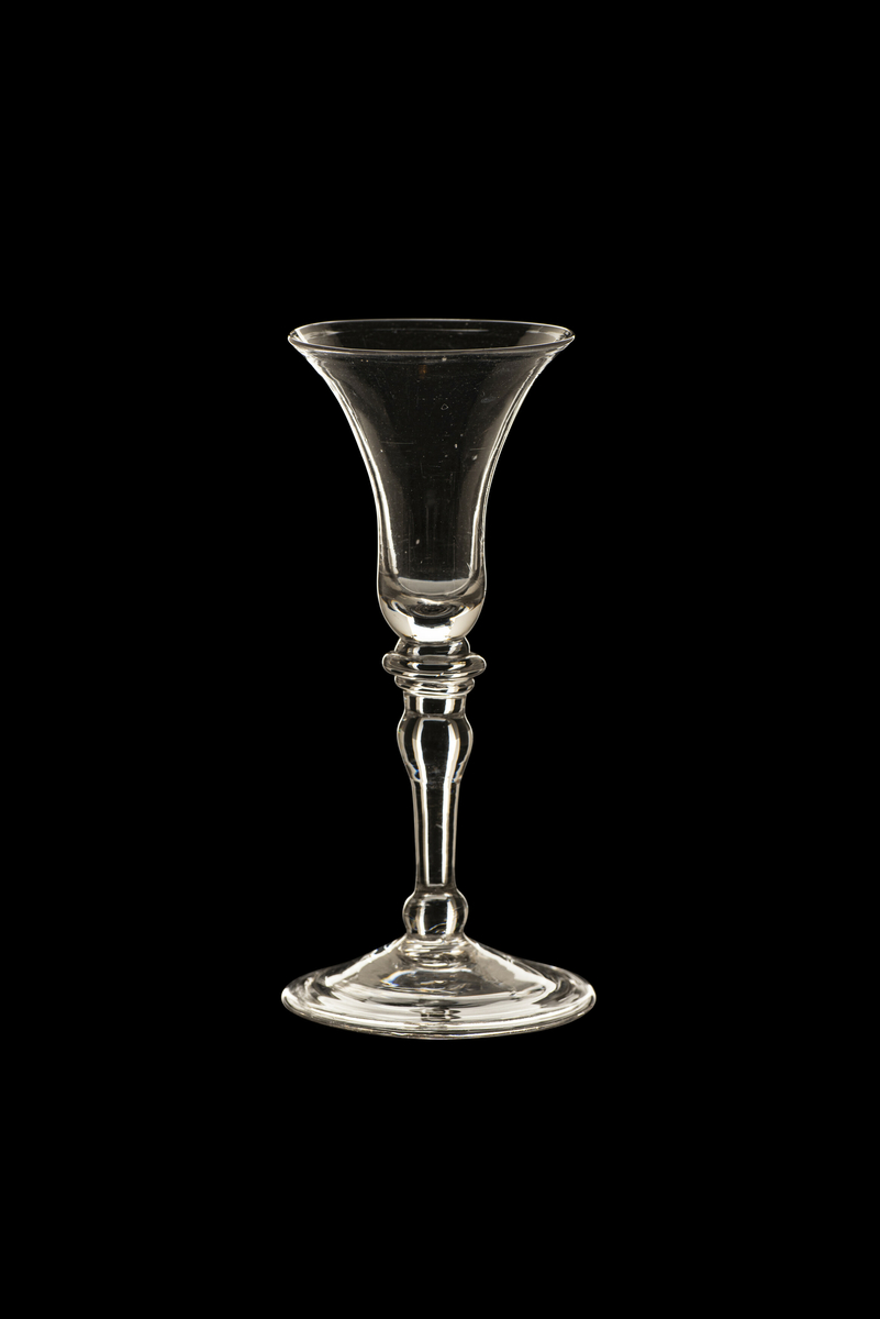 Brennevinsglass, variant av "Trundhiems Brendevins Spits Glas". Rent, tilnærmet fargeløst glass. Trompetformet klokke med slank, balusterformet stett. To skiver under klokken og kuler i stettens ender. Ombøyet fotkant. 