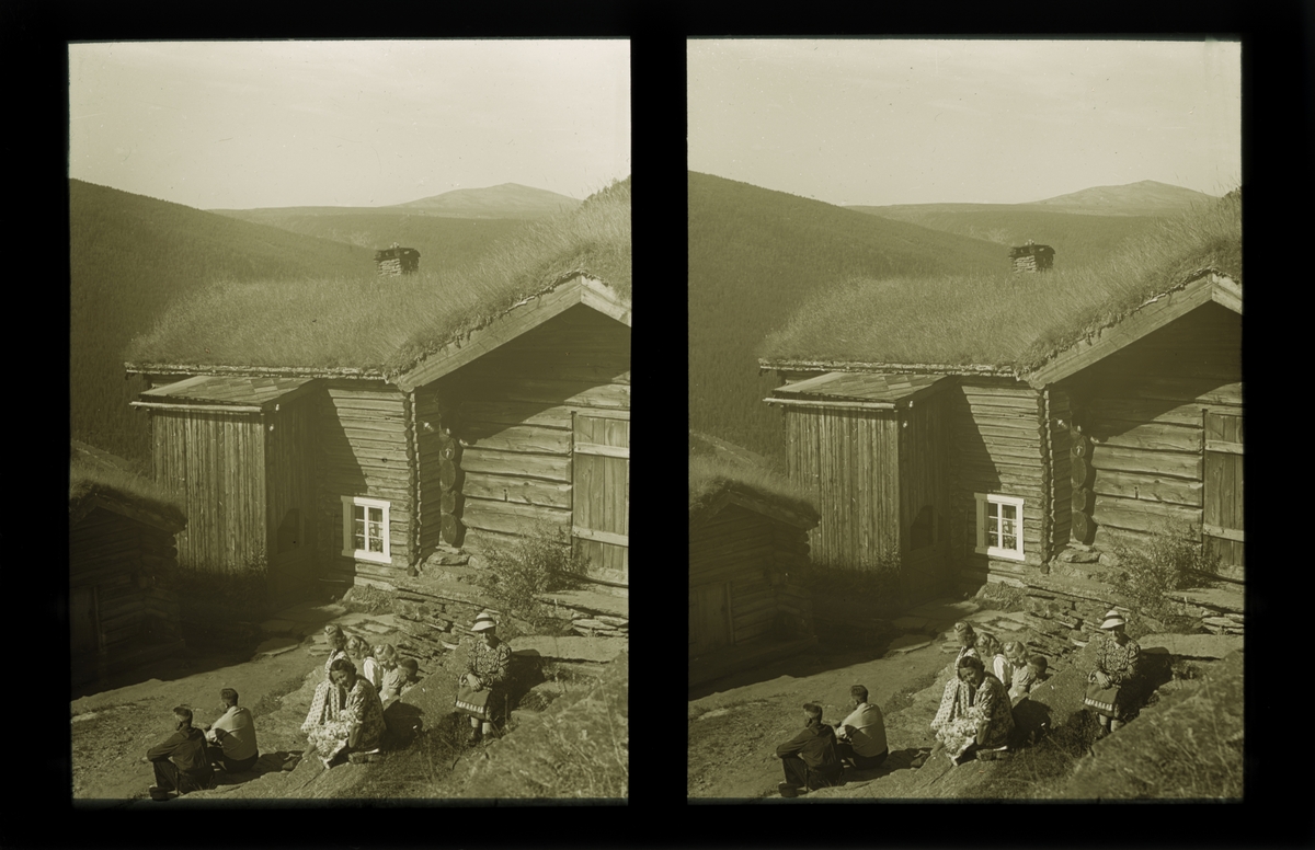 Ene hjørnet av en laftet stue. På tunet sitter en gruppe kvinner og menn og slapper av. Fjelllandskapet viser i bakgrunnen. Tilhører Arkitekt Hans Grendahls samling av stereobilder.