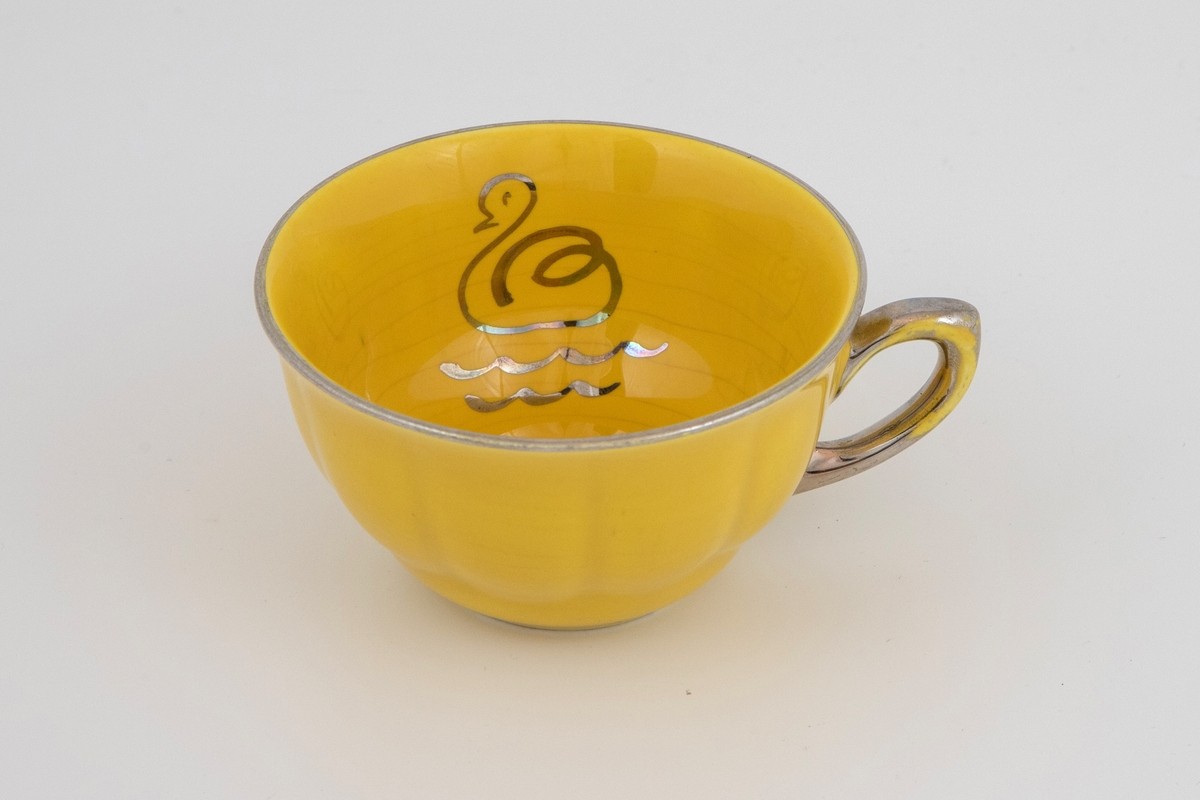 Sterkt gul kopp, med sølv dekor i form av and/svane på 2 bølgelinjer innvendig i koppen. Også sølv langs munningen og på hanken. Uklart om det dreier seg om kaffekopp eller en liten tekopp. Rund bolleform, som minner om puddingform med sin buklete ytterside. Avsmalende nederst, med antydning av stett.