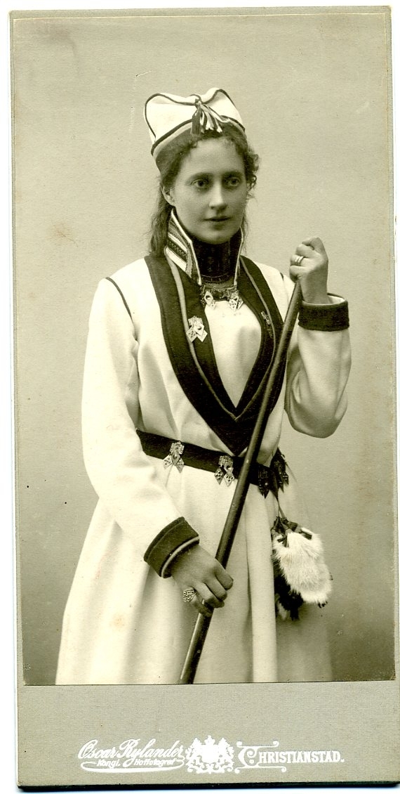 Kabinettsfotografi: porträtt av okänd ung kvinna i en variant av folkdräkt som för tankarna till samedräkt. Hon håller en vandringsstav i händerna.