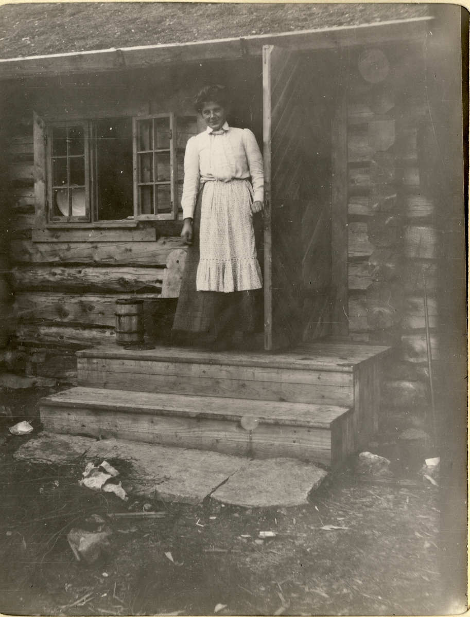Tjenestepiken Adolphine på trappen til jakthytte ved Fjergen i Meråker, tilhørende Nils O. Y. Fearnley. Antagelig fotografert 1908.