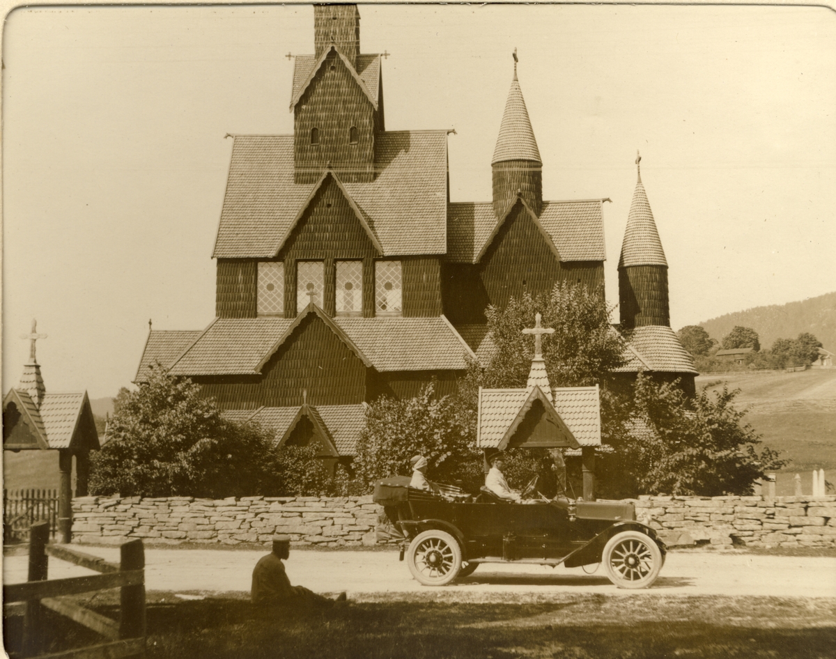 Nini Egeberg og familiens sjåfør fotografert i bilen foran Heddal stavkirke. I forgrunnen sitter en mann på plenen. Fotografert 1913.