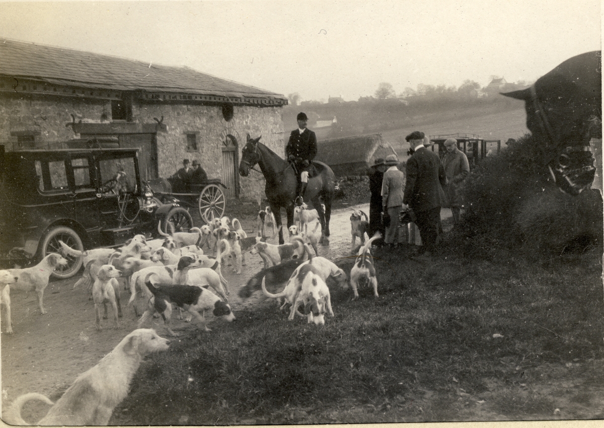 En jeger til hest og hunder fra "The Monmouthshire Hounds" på vei til eller fra jakten "The Meet". Til venstre foran driftsbygning en bil og et hestekjøretøy med en kvinne og en mann. Til høyre er to kvinner og to menn tilskuere. Antagelig fotografert på Coldbrook Park i Abergavenny, Wales i 1913.