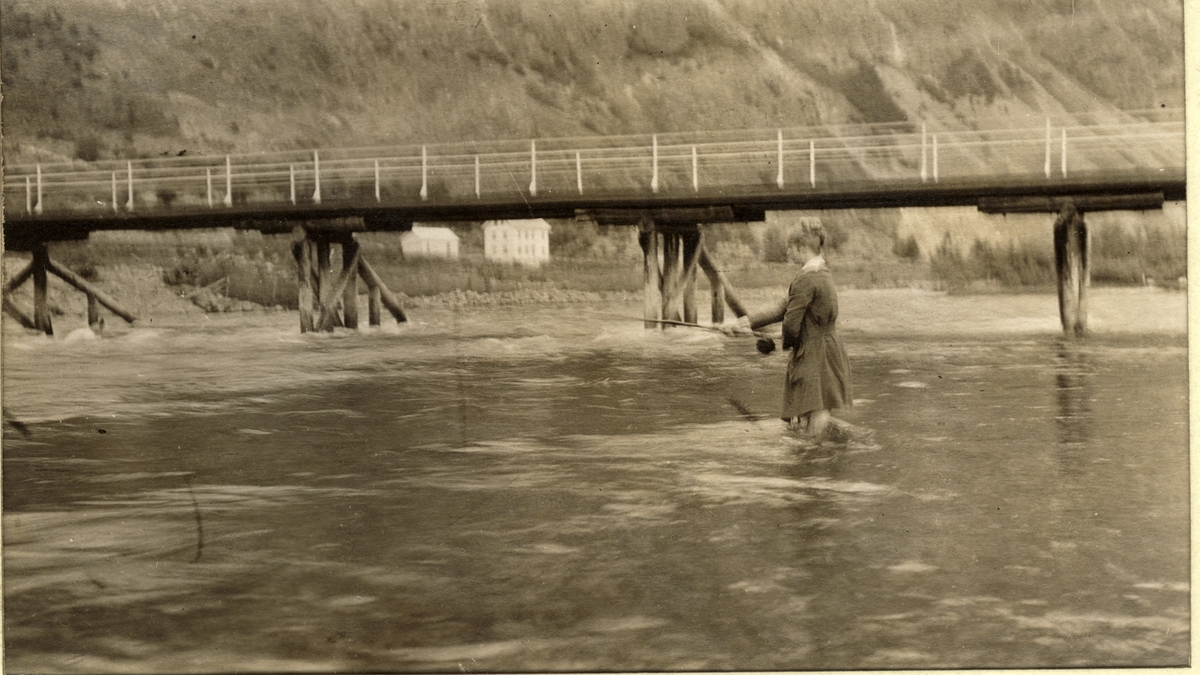 Evelyn Egeberg fisker i Lærdalselvi, Sogn og Fjordane. Bro over elven og våningshus med uthus i bakgrunnen. Fotografert sommeren 1916.