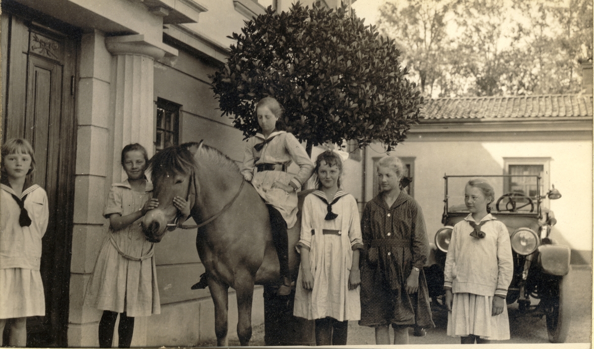Pinse på Bogstad gård. Seks jenter, hvorav én til hest, ved inngangspartiet på gårdsplassen. I bakgrunnen står en bil. Fotografert 1919.