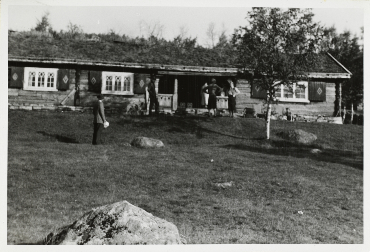 To kvinner og to menn skimtes foran hytten til fru Lizzie Michelet på Hovden i Møsstrand, Vinje, Telemark.