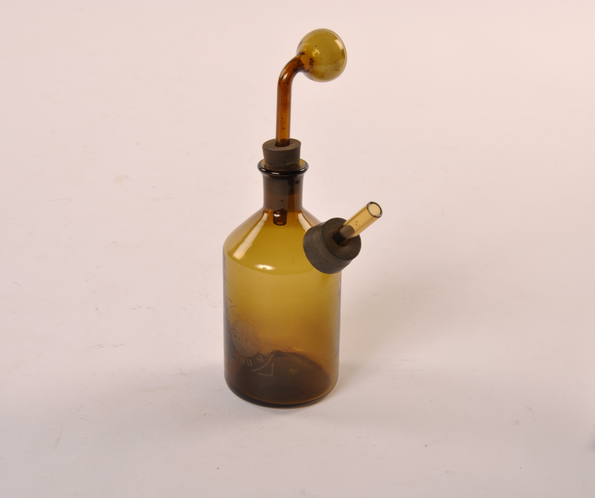Flaske med avsmalet tut og en avsmalet utstiikkende åpen sylinder. Løs bøyd sylinder festet med gummipropp. Flasken er i brunt glass.