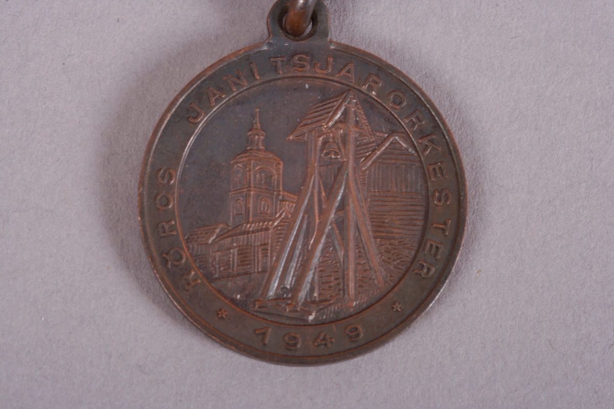 Medalje med påskrift Røros janitsjarorkester, 1949.
På forsiden motiv fra Røros sentrum med klokke og kirketårn.

På baksiden påskrevet: Trøndelag Hornmuiskkforbund 16 & 17 juli.