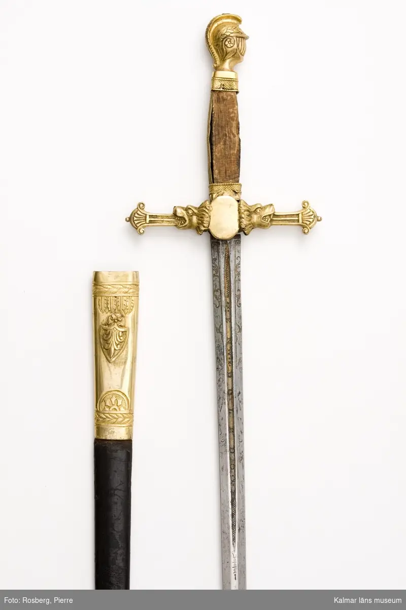 KLM 14188. Ämbetsvärja med gehäng. Med toledoklinga. Rak tveegggad klinga med blodrand. I och vid blodranden etsade och förgyllda ornament samt inskriften Sebastian Ernandes En Doledo. Korsfäste av förgylld mässing, parerstången i form av varghuvuden med palmetter i gapen. Knapp i form av antik hjälm. Fyrsidig kavel, som på båda sidorna varit klädd med pärlemorplattor. Balja (ej komplett, nedre delen av baljan saknas) av lackskinn med munbleck av förgylld mässing. Hake i form av trekantig sköld. Värjgehäng av blått kläde med sölja och spänne av förgylld mässing, i spännet trekronors vapen.