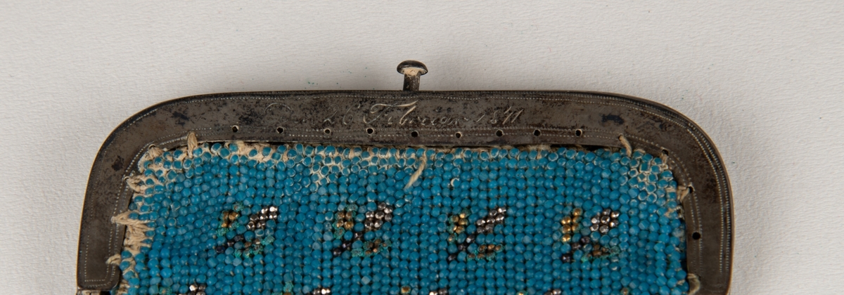 Liten pung, med avrundede nedre hjørner, med  små perler i egyptisk blått og gyllent, og mørkebrunt, siksakbord nederst. På spennen   inngravert  :   E J C L den 26 Februar 1841