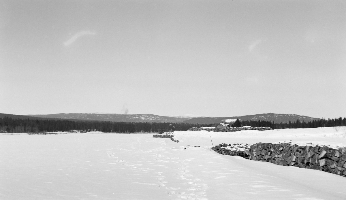 Vassdragsvesenets forbygninger mot isgang ved Alme i Åmot, fotografert vinteren 1933.  Fotografiet er tatt fra ei flat, snødekt slette, antakelig isen på Glomma.  Til høyre i bildet ses et slags steingjerde, som skal være opplagt som en forbygning mot blant annet isgang i elva.  Deler av forbygningen er dekt av snø og is, noe som i underteksten under albumkopien av fotografiet tas som et tegn på at muren er for lav.  Bak muren, om lag midt på bildet, bak en......, skimtes takene på bygningene på gardsbruket Alme.  I bakgrunnen en lave åser med barskog. 