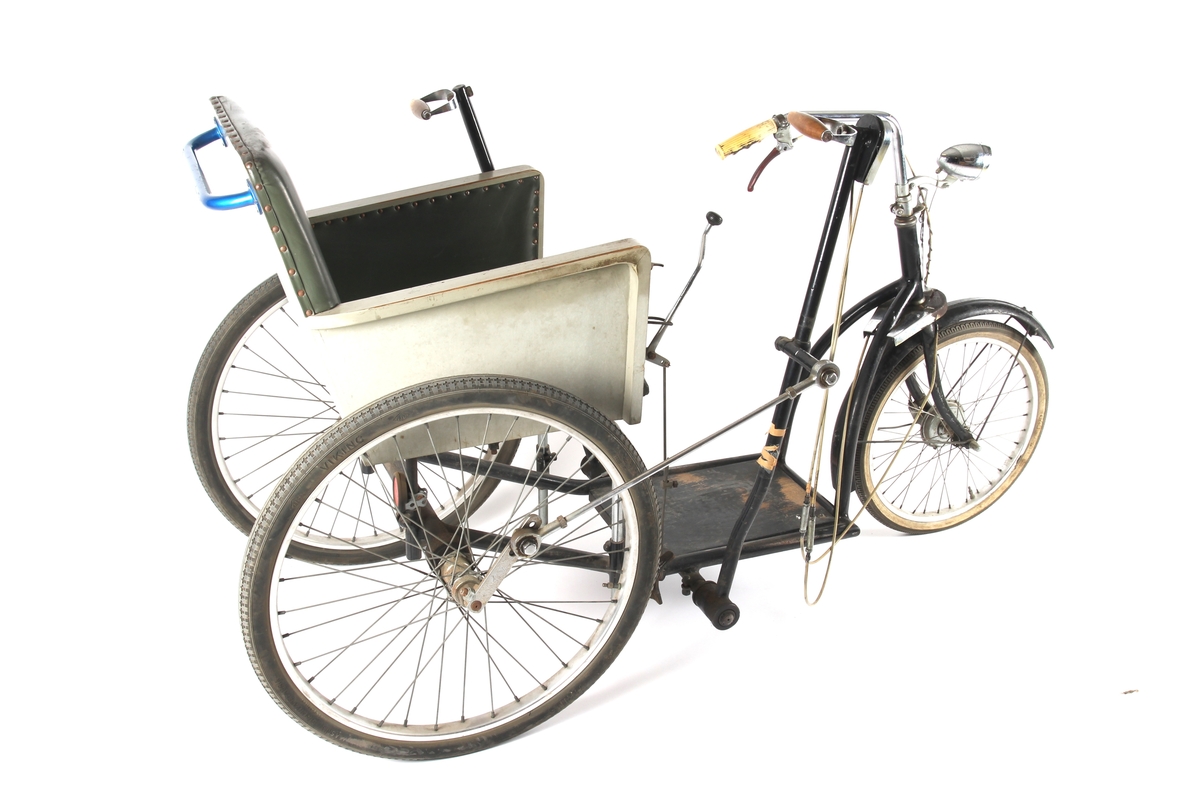 Sortlakkert invalidvogn på tre hjul med benstøttebrett og stol. Trommelbremsenav på forhjulet med bremsegrep på styrestangen. Sykkelen er hånddrevet.
