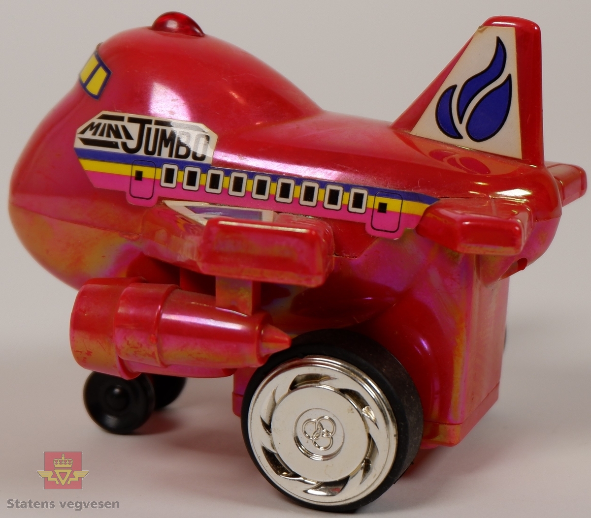 Miniatyrfly i fargen rød med klistremerker som har påskriften MINI JUMBO. Den er utstyrt med en mekaniske som gir den fremdrift når man trekker hjulene bakover.