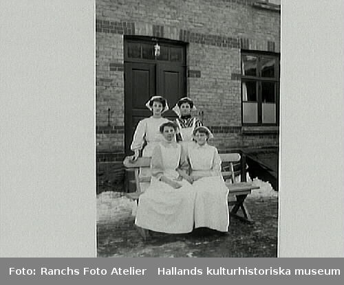 Fyra flickor ur personalen på Knutssons Marmeladfabrik, fotograferade vid bänk utomhus med snö längs fasaden. Fabriken låg på Drottninggatan 5, kv Flickskolan 6, Varberg.