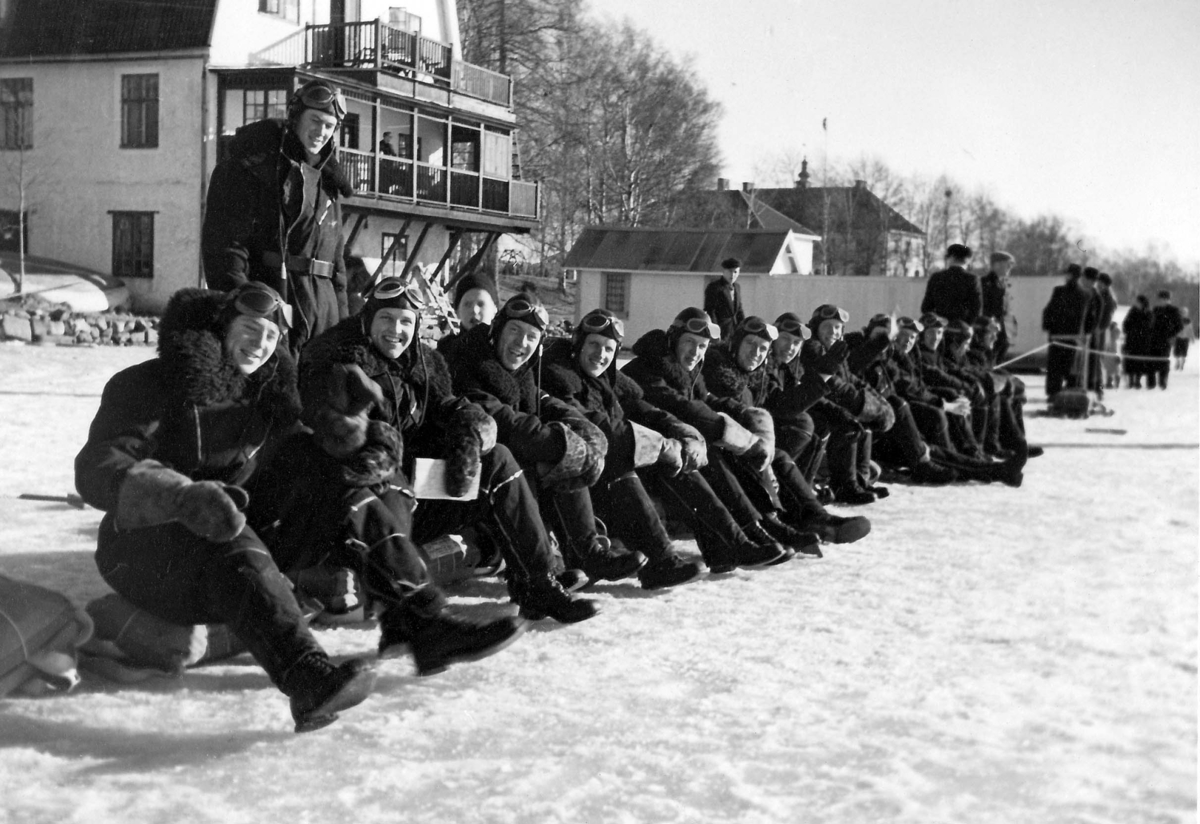 Grupporträtt av militära flygare i flygoveraller under rast, sittandes framför byggnad, vintertid. 1930-tal.