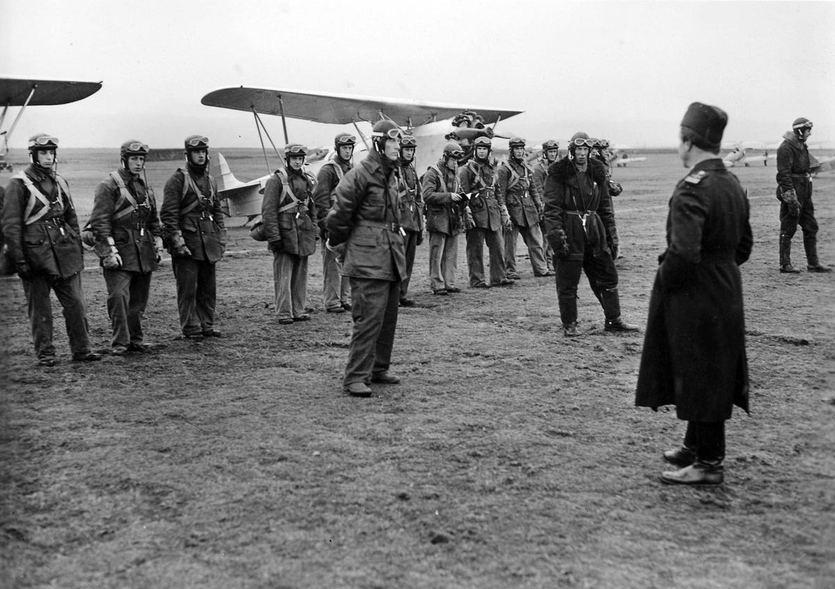 Nils Söderberg ger militära flygare order inför flygning, under 1930-talet. Omkring 15 flygare samlade på ett flygfält framför flygplan S 6. 1930-tal.