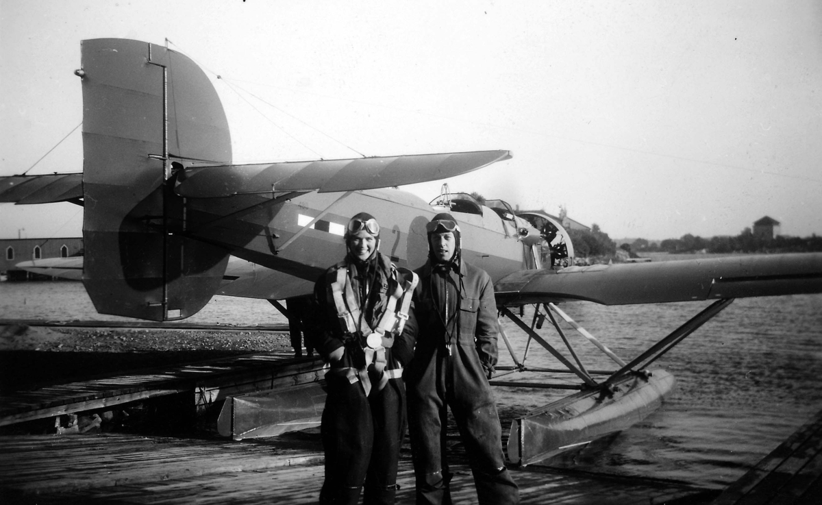 Gunborg och Verner Tornberg står iklädda flygdräkt på brygga framför flygplan S 5C nr 480 som står på vatten. Kronmärket är övermålat. 1930-tal.