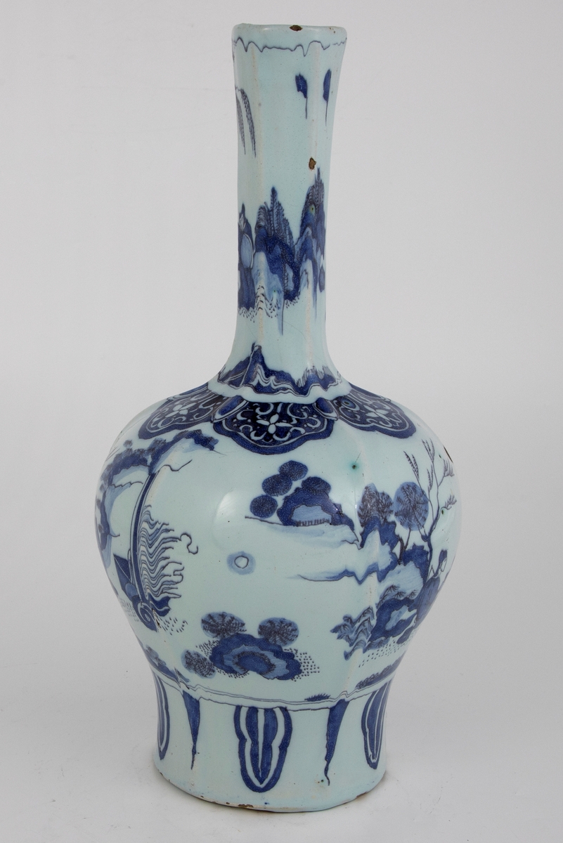 Heksagonal klubbeformet vase med kinesiskinspirert motiv med figur i landskap på både korpus og vasehals. En europeisk versjon av lingzhi/ ruyi-ornamentikk rundt vasehalsen på toppen av korpus.  Palmeblader nederst på vasen under hovedmotivet.