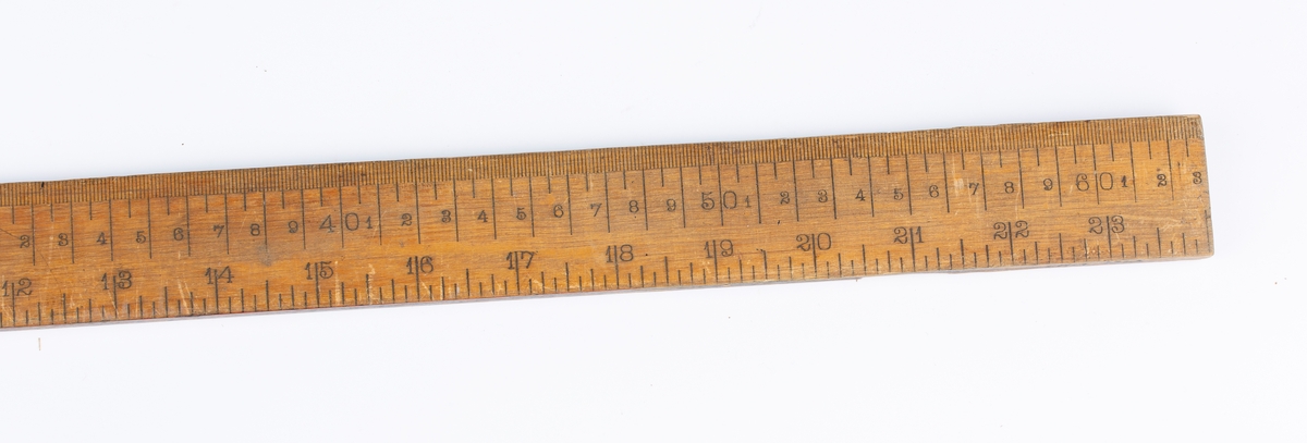Flat, smal målestav med inndeling i cm og tommer. Lengden er lik én alen.