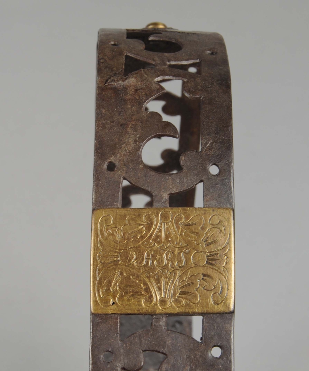U-formet klave av metall med akantusmønster og en krok i den ene enden. Det er to messingskilt festet på klaven og en messingblomst på midten.