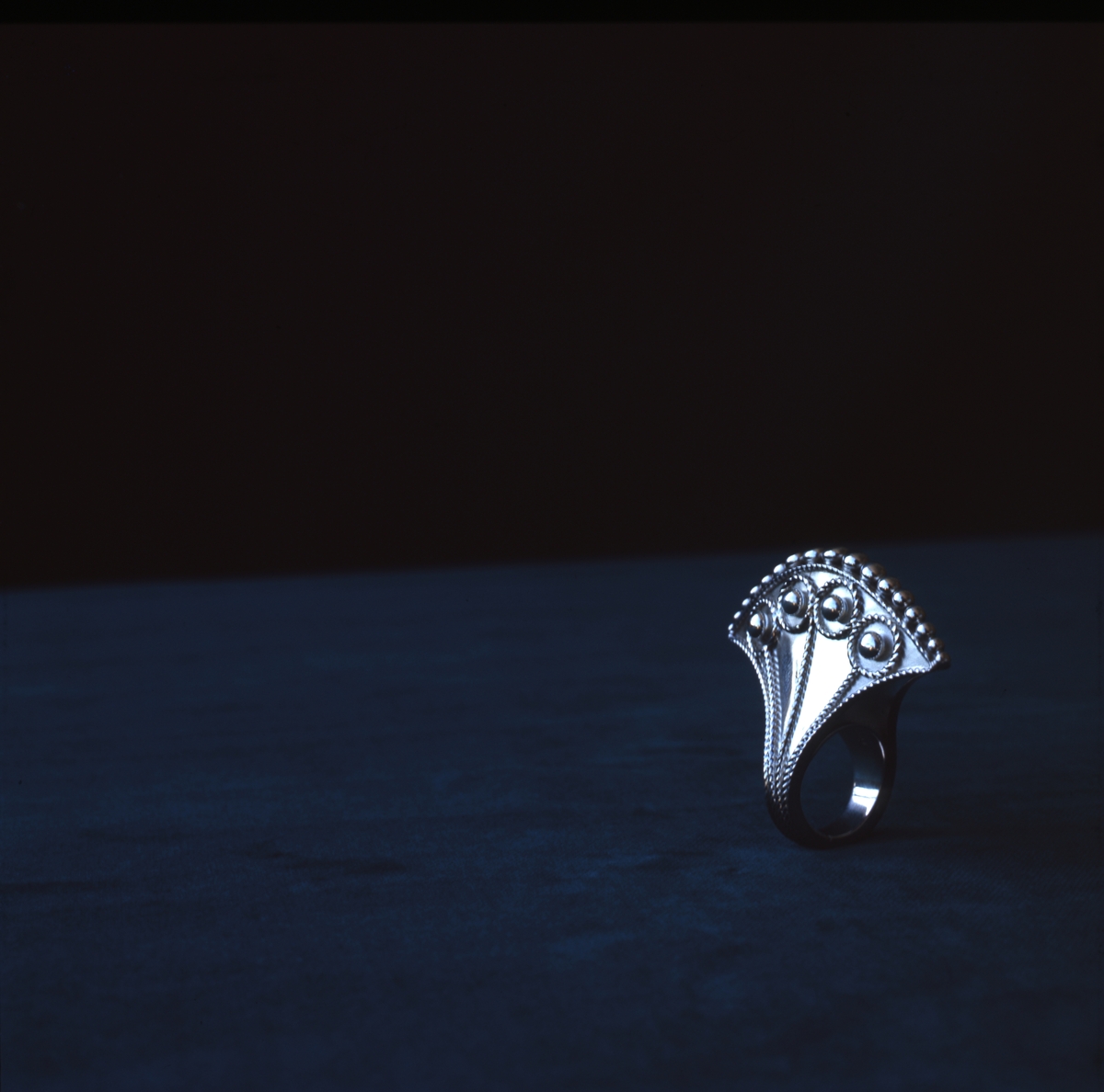 Silverring tillverkad av Rosa Taikon fotograferad mot mörk bakgrund. Ringen består av silverplåt formad som en yxa, en form som Taikon ofta använder i sina verk. Ringen är dekorerad med filigrantråd och granuler.