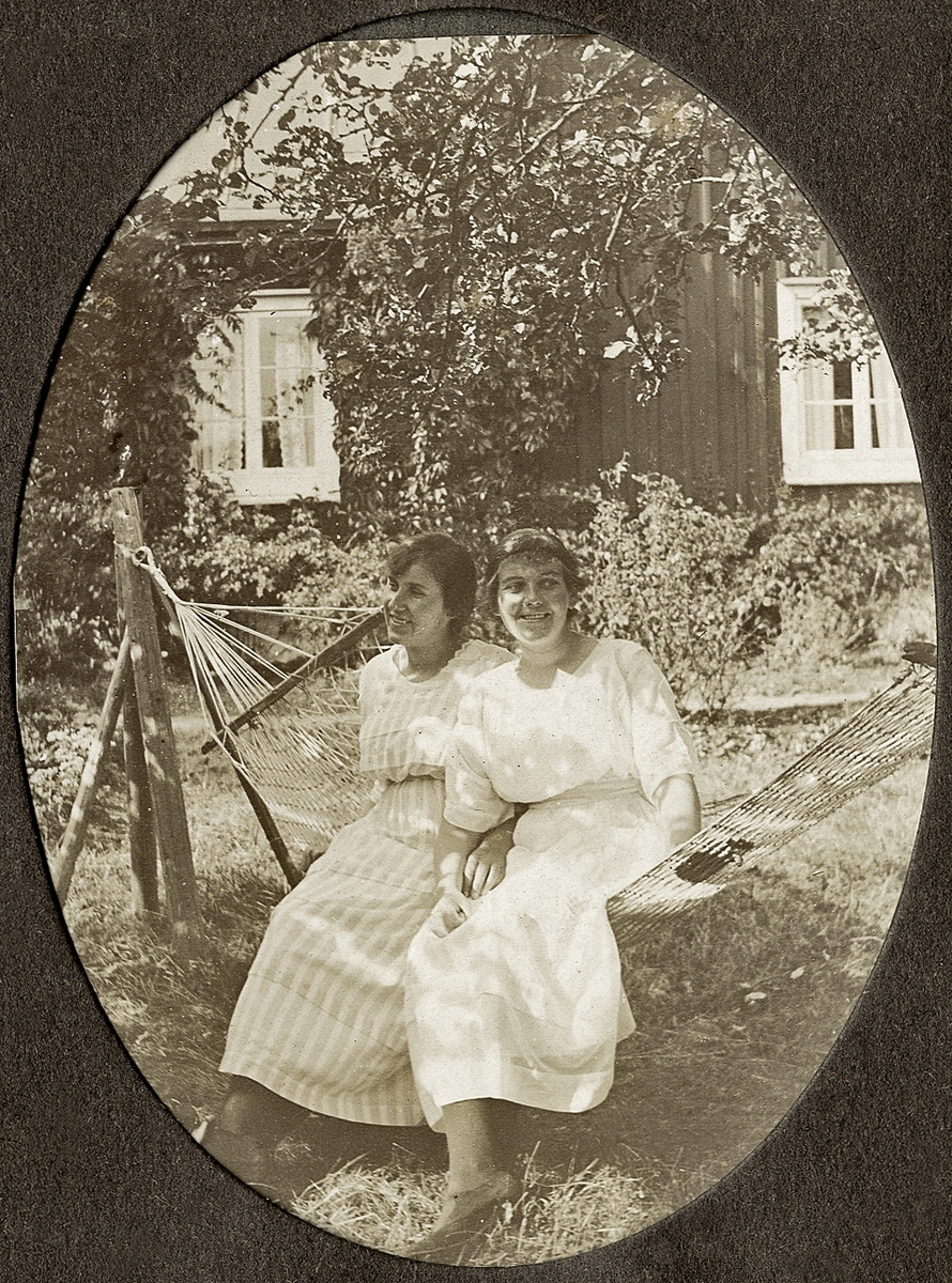 Två kvinnor sitter i en hängmatta i en lummig trädgård. I bakgrunden syns ett bostadshus.