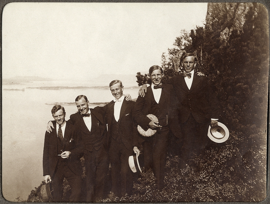 Fem glada unga män står uppradade på en höjd vid havet.