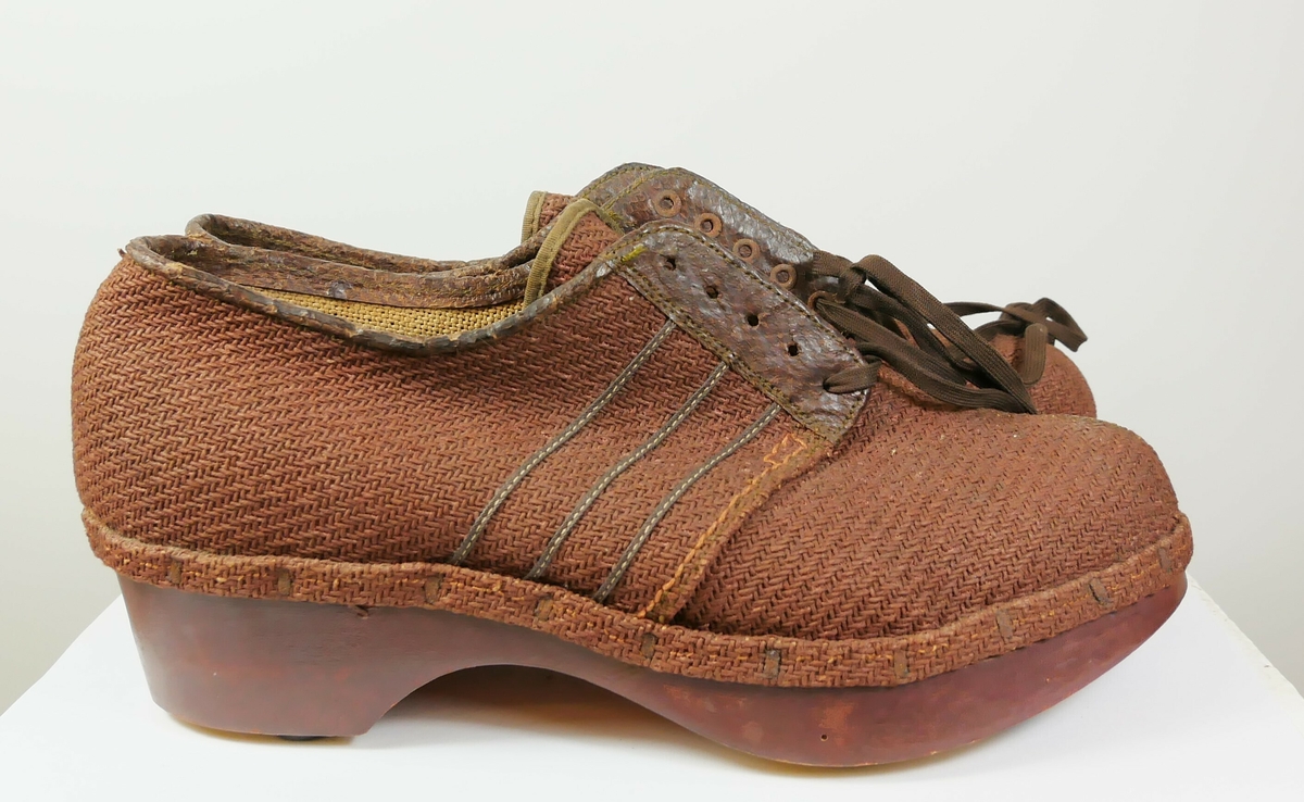 Røde sko av tekstil som er naglet til en en rødmalt såle av tre Under sålen er det ni gummiknotter. Det er brune lisser til knyting foran på skoen. 