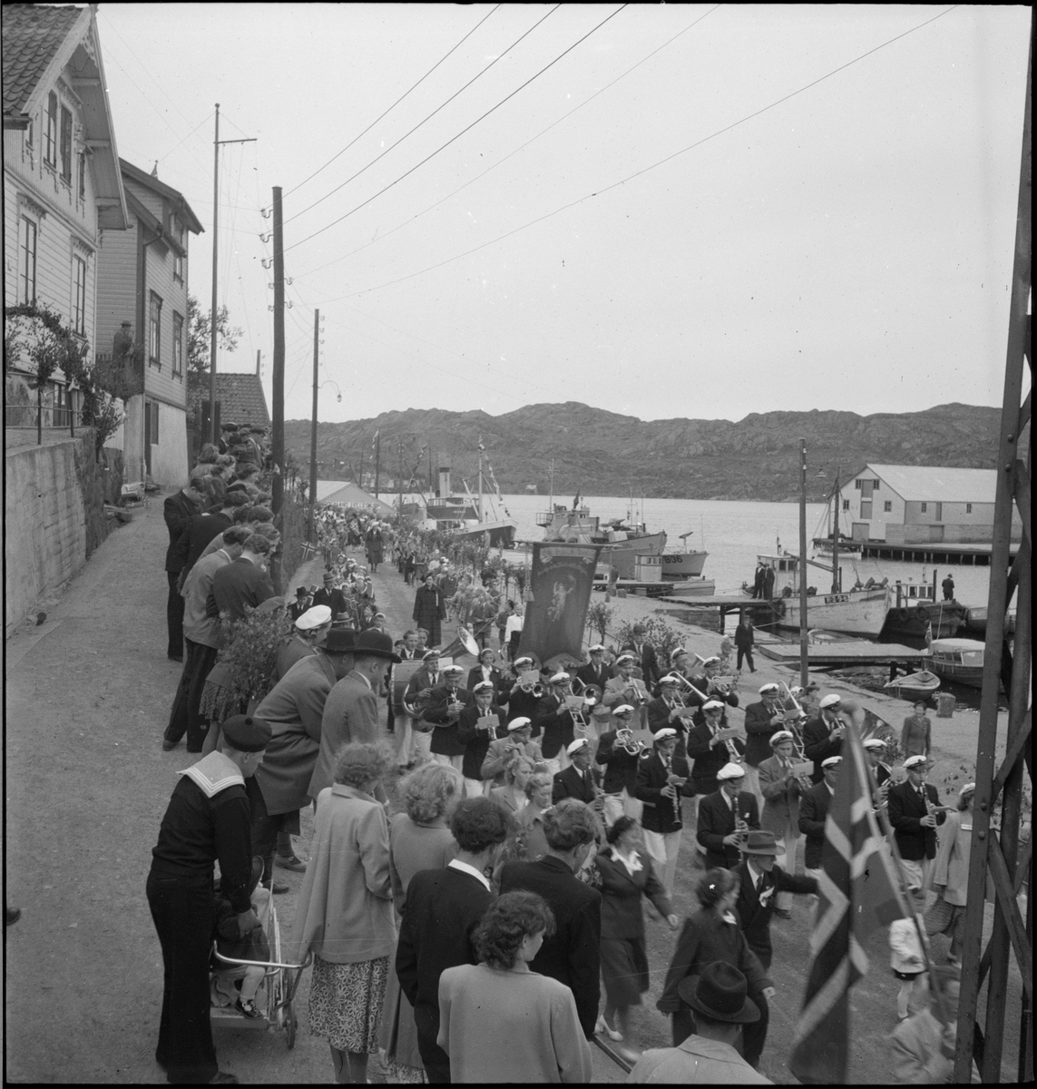 Folketog på 17. mai. Flere korps og barn går langs Vågen og igjennom sentrum av Egersund. Mange har kommet for å se toget.