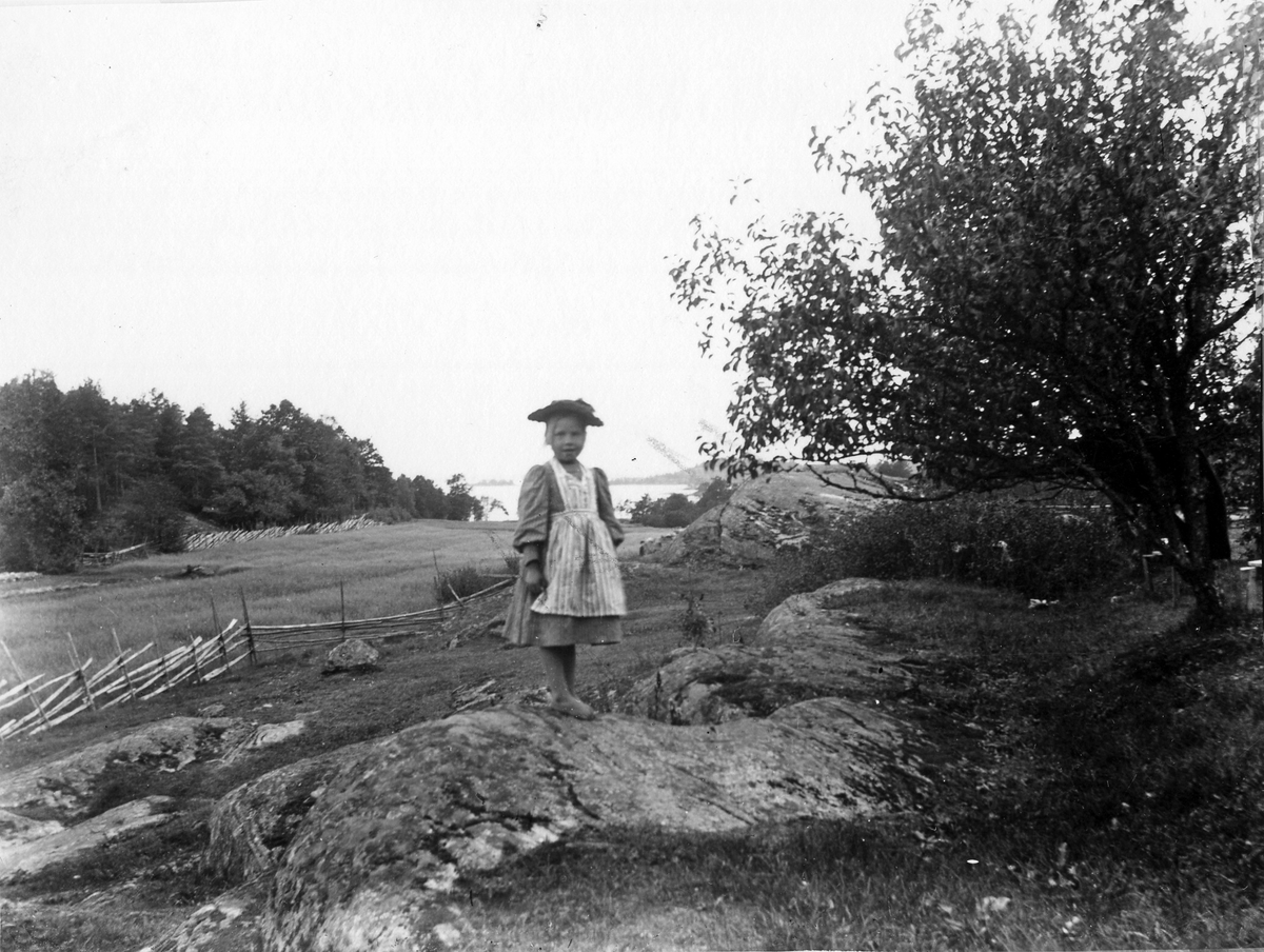 En flicka i klänning, förkläde och hatt står på en sten.
