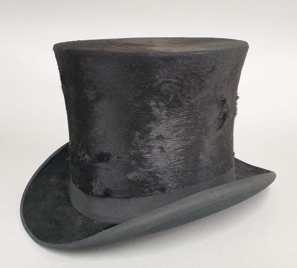 Flosshatt med hatteeske. Hatten har ripsbånd rundt pullen og flosset overflate. Esken har håndtak og fester av metall, og stempel på den ene kortsiden.